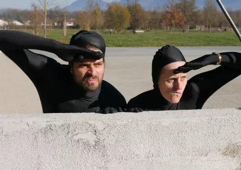 un homme et une femme en combinaison noire et gantés de noir, cachés derrière un mur une main en visiere sur leur fond © Ville de Crolles